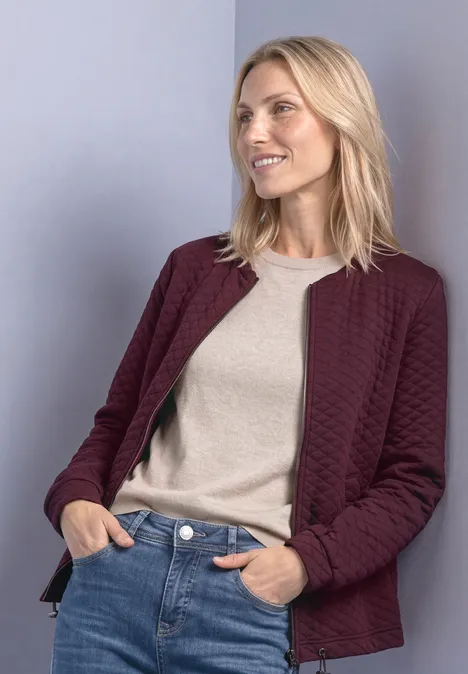 Jasje met ronde hals en structuur rustic rouge Blonde vrouw in bordeaux doorgestikte jas, licht shirt en jeans, glimlachend opzij kijkend.