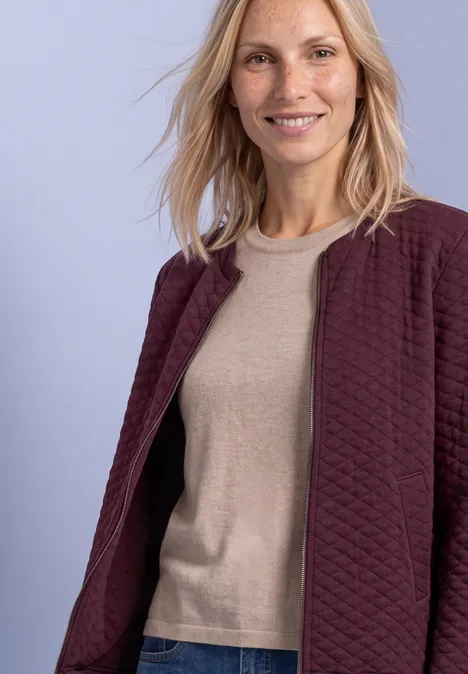 Jasje met ronde hals en structuur rustic rouge Lachende vrouw met blond haar draagt een bordeaux gewatteerd jasje en beige trui voor een pastelkleurige achtergrond.