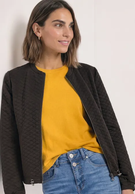Jasje met ronde hals en structuur black coffee Vrouw met een zwarte doorgestikte jas, een geel shirt en blauwe jeans, licht glimlachend.