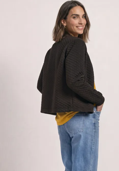Jasje met ronde hals en structuur black coffee Vrouw in zwarte, gestructureerde jas, geel shirt en spijkerbroek kijkt lachend over haar schouder.