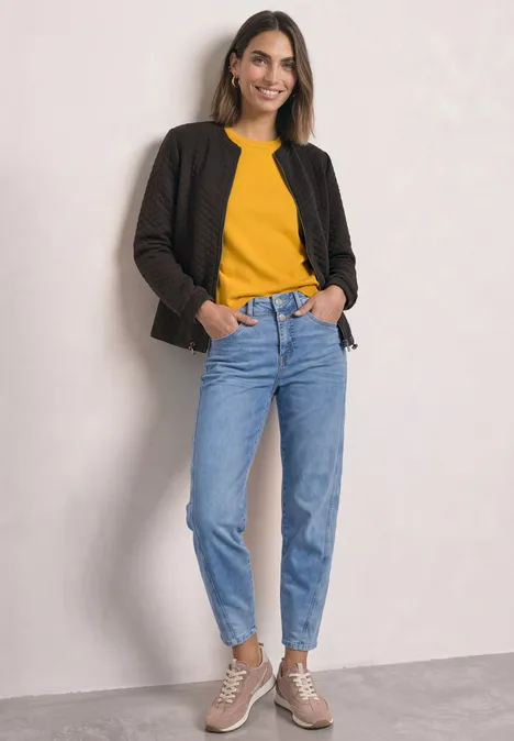 Jasje met ronde hals en structuur black coffee Een vrouw glimlacht en draagt een geel shirt, zwarte jas, blauwe jeans en beige sneakers voor een lichte muur.