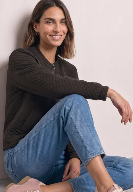 Jasje met ronde hals en structuur black coffee Jonge vrouw zit relaxt op de grond, draagt een jeans en een donker sweater en lacht ontspannen.
