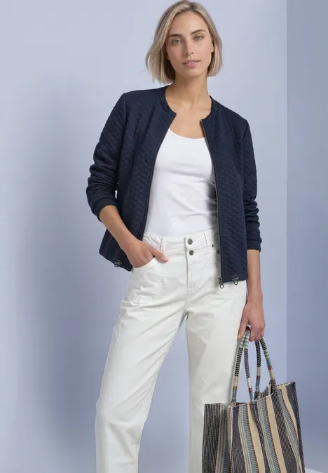 Frau trägt weiße Hose, weißes Top, dunkelblaue gesteppte Jacke und eine gestreifte Stofftasche.