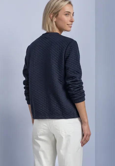 Frau steht seitlich, trägt dunkelblaue gesteppte Jacke und weiße Hose vor neutralem Hintergrund.