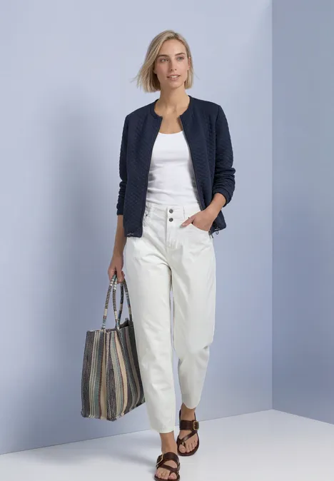 Frau in weißer Hose und Top mit dunkelblauer Strickjacke, gestreifter Tasche und Sandalen vor neutralem Hintergrund.