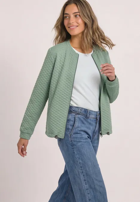 Eine lächelnde Frau trägt eine grüne Steppjacke, weißes Shirt und Jeans.