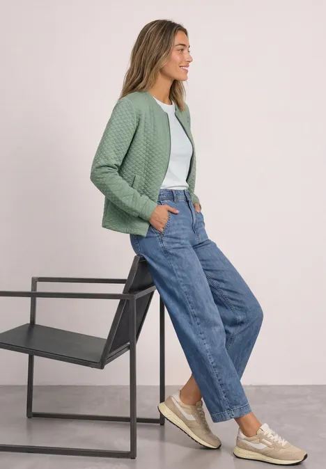 Frau lehnt an schwarzem Stuhl, trägt grüne Strickjacke, weißes Shirt, weite Jeans und beige Sneaker.