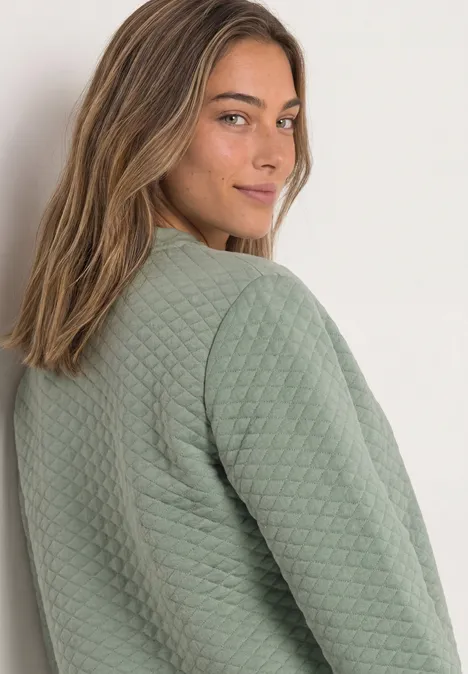 Eine Frau in einem gesteppten, grünen Pullover blickt über die Schulter, leicht lächelnd.
