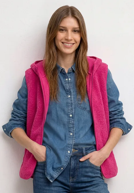 Jonge vrouw glimlacht in spijkerblouse en roze fleecevest voor een neutrale achtergrond.