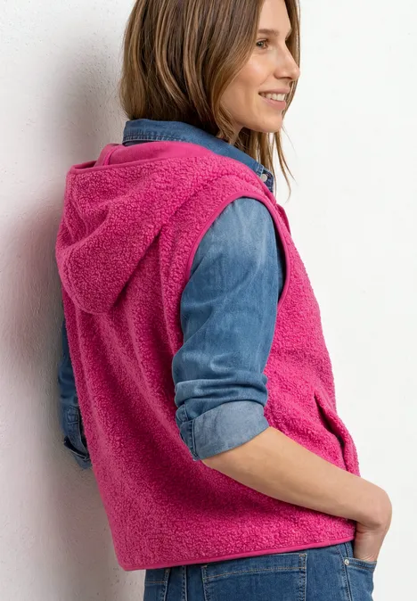 Vrouw draagt roze, zachte hoodie-vest over denimhemd, staat zijwaarts en glimlacht.