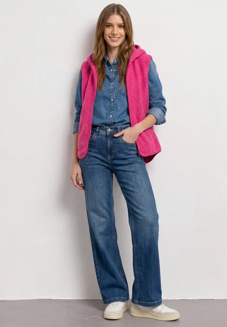 Vrouw in denimblouse en -jeans, roze gewatteerd gilet en witte sneakers, handen in zakken.