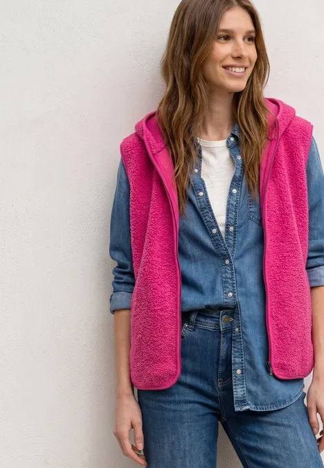 Een glimlachende vrouw draagt een blauwe denimblouse en jeans met een roze mouwloze fleecebodywarmer.