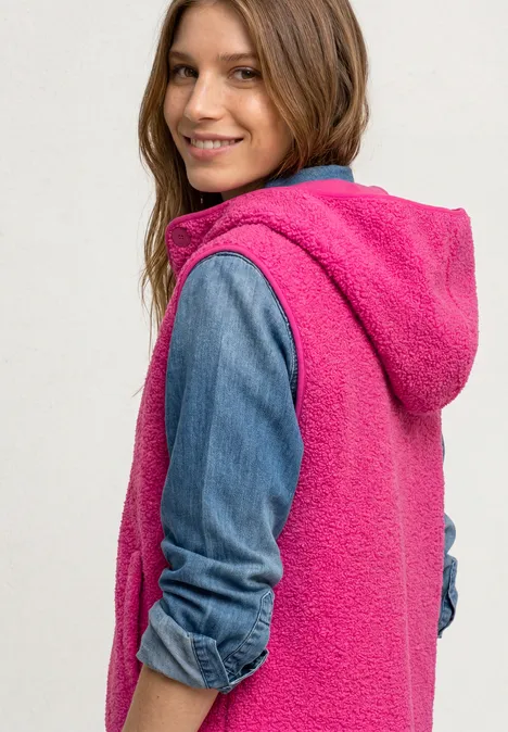 Vrouw in een roze fleece-vest met capuchon over een spijkerblouse, glimlachend over haar schouder kijkend.