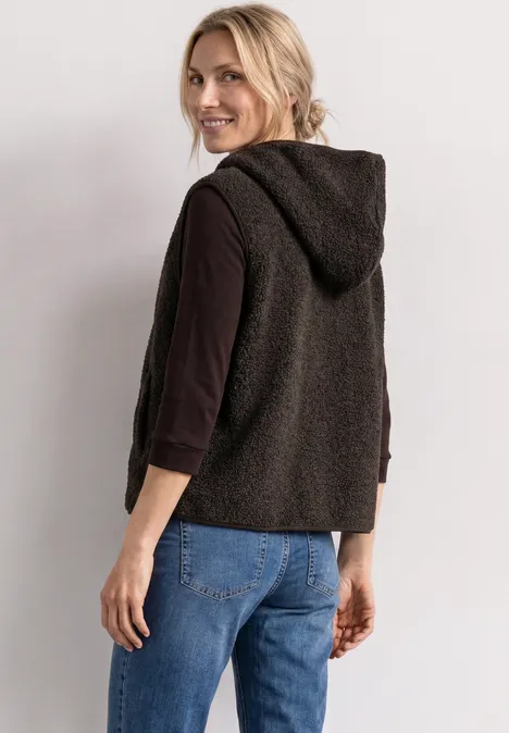 Teddy-Weste mit Kapuze und Zipper black coffee Eine Frau trägt eine schwarze Kapuzenweste über einem dunklen Langarmshirt und Jeans, schaut über die Schulter.