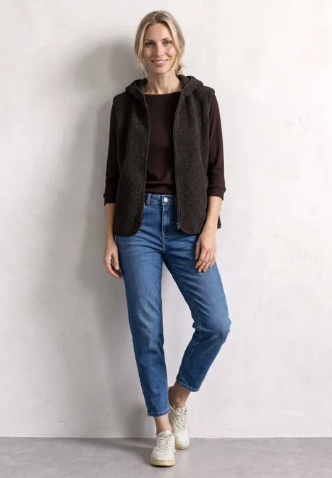 Teddy-Weste mit Kapuze und Zipper black coffee Junge Frau steht vor heller Wand, trägt braune Jacke mit Kapuze, braunes Shirt, blaue Jeans und weiße Sneakers.