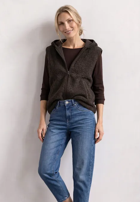 Teddy-Weste mit Kapuze und Zipper black coffee Eine Frau trägt eine braune Kapuzenweste über dunkelgrauem Shirt und blauen Jeans vor heller Wand.