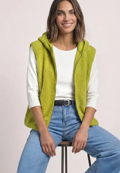 Vrouw zit op kruk, draagt groene fleecevest met capuchon, wit shirt en blauwe jeans, lacht.