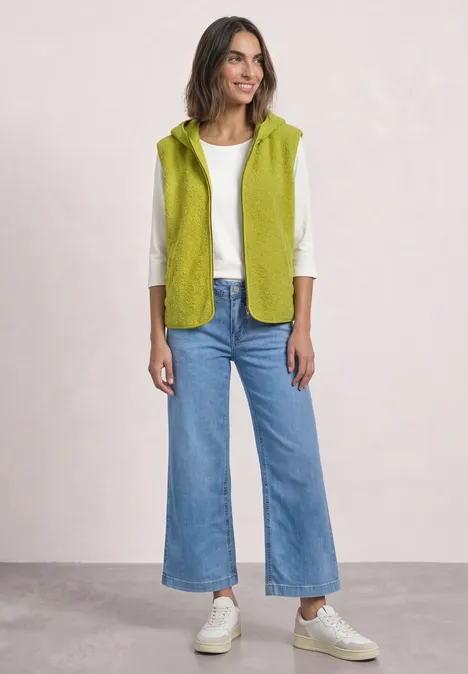 Vrouw in lichtgroene mouwloze bodywarmer, wit shirt met lange mouwen, wijde jeans en sneakers.