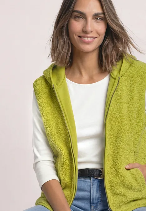 Glunderende vrouw in een groene fleecebodywarmer met capuchon, wit shirt en blauwe jeans.