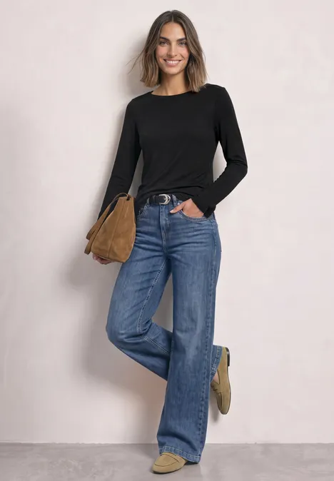 Eine lächelnde Frau in schwarzem Oberteil, weiter Jeans und brauner Tasche vor heller Wand.