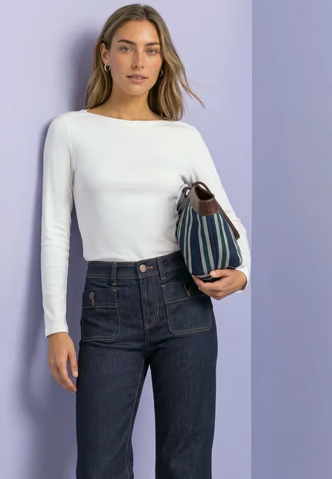 Bluzka Basic z dekoltem w łódkę off white Kobieta w białej bluzce z długim rękawem i ciemnych dżinsach trzyma pasiastą torebkę na fioletowym tle.
