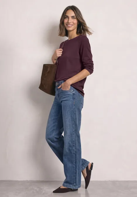 Frau in lässigem Look mit weiten Jeans, bordeauxfarbenem Pullover und brauner Tasche.