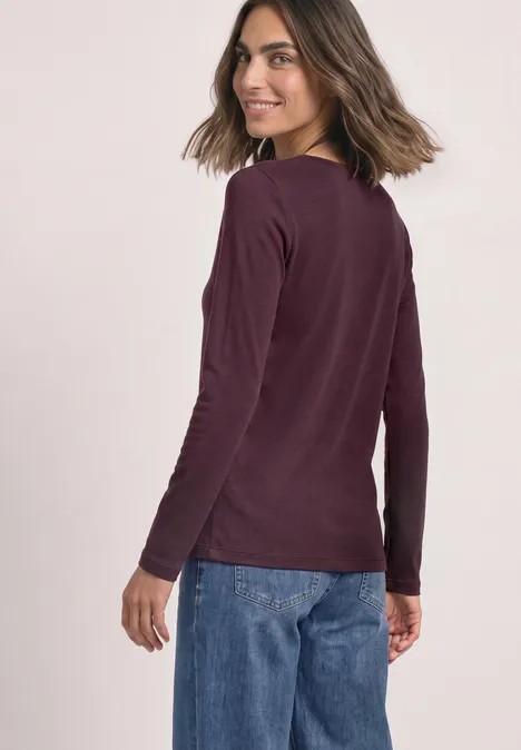 Eine Frau trägt ein bordeauxrotes Langarmshirt und blaue Jeans, blickt über die Schulter zur Kamera.