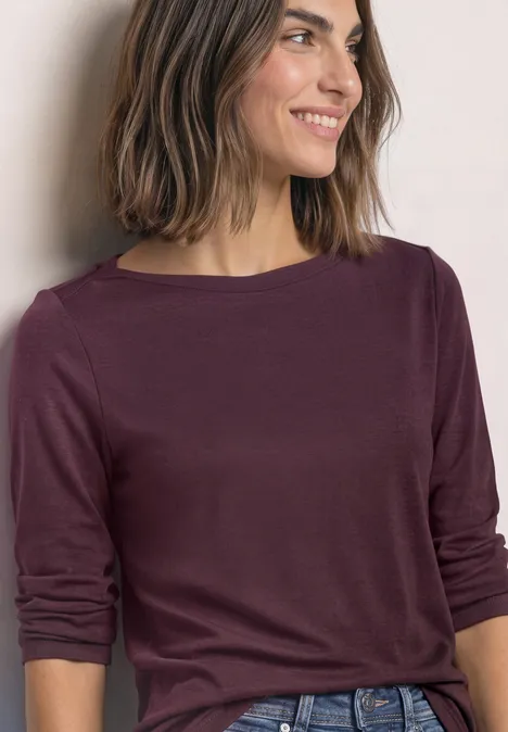 Frau mit schulterlangen Haaren in weinrotem Langarmshirt, lächelt seitlich.