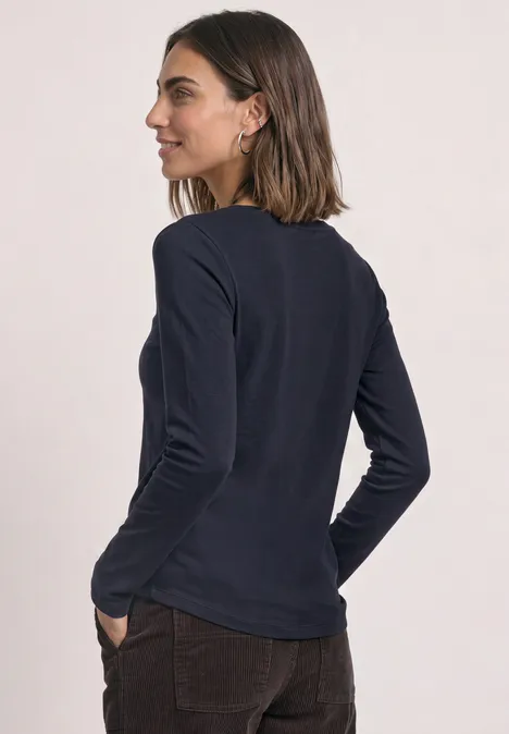 Eine Frau im Profil trägt ein dunkelblaues Langarmshirt und braune Hose, Hände in den Taschen.