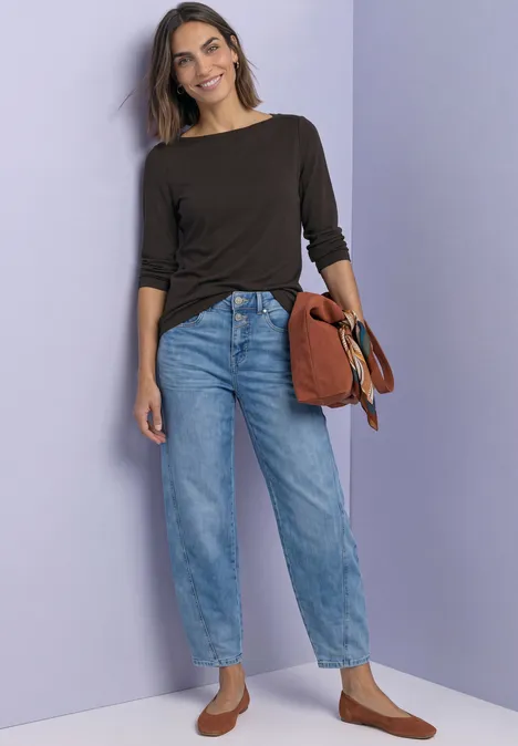 T-shirt basique à col bateau black coffee Frau vor lila Wand trägt dunkelbraunes Oberteil, helle Jeans, braune Schuhe und hält eine braune Tasche.