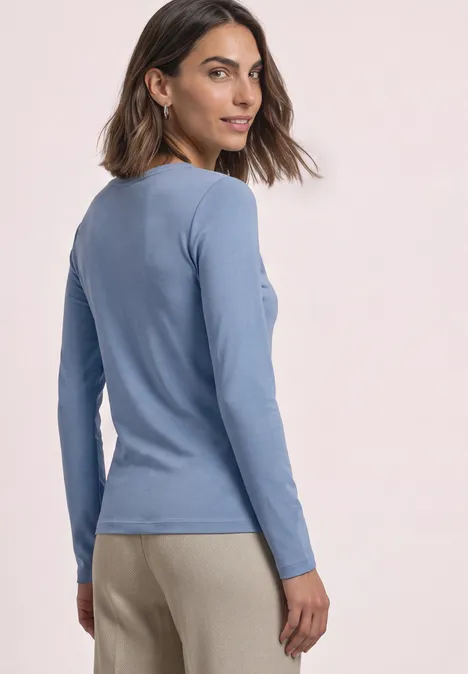 T-shirt basique à col bateau northern blue Eine Frau in blauem Langarmshirt und beigen Hosen schaut über die Schulter, vor hellem Hintergrund.