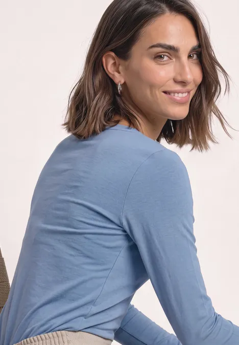 T-shirt basique à col bateau northern blue Eine lächelnde Frau in einem blauen Langarmshirt schaut über ihre Schulter.