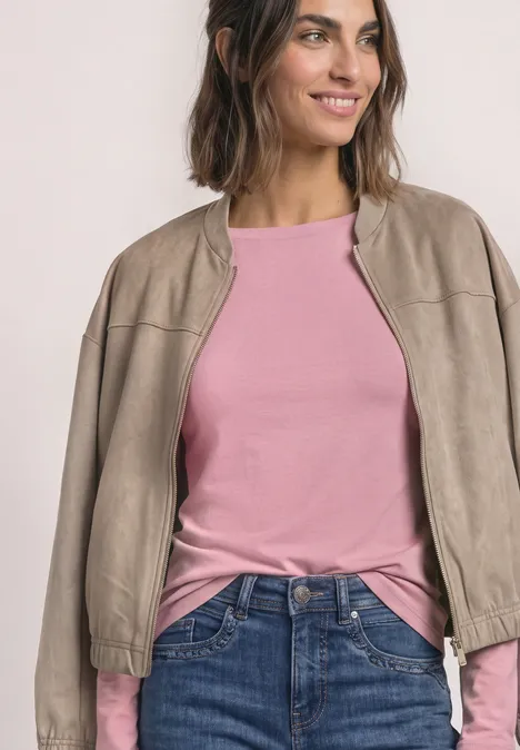 Een glimlachende vrouw in een roze longsleeve, beige leren jack en spijkerbroek voor een lichte achtergrond.