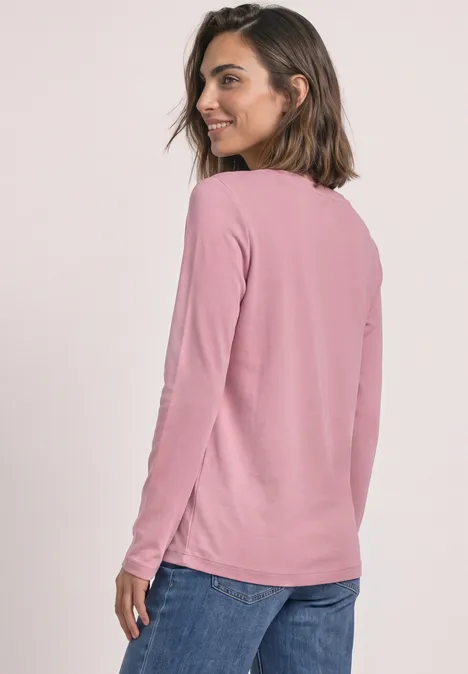 Een vrouw draagt een roze longsleeve en een blauwe jeans, lacht terwijl ze over haar schouder kijkt.