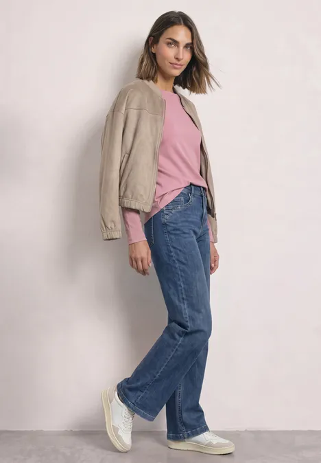 Een vrouw leunt tegen een muur, draagt een beige blazer over een roze T-shirt, wijde spijkerbroek en witte sneakers.