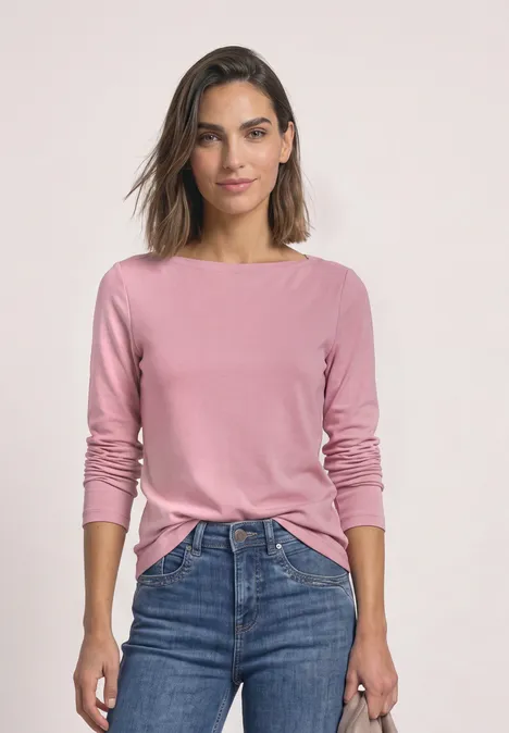 Vrouw in een roze longsleeve en blauwe jeans, staand voor een neutrale achtergrond.