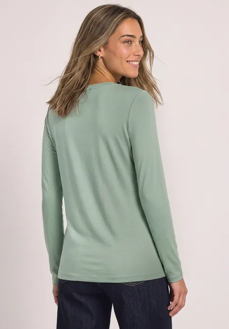 Frau im mintgrünen Langarmshirt und Jeans, Blick seitlich über die Schulter.