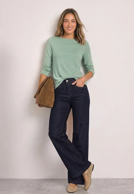 Eine lächelnde Frau trägt eine mintgrüne Bluse, weite Jeans und flache Schuhe, hält eine braune Tasche.