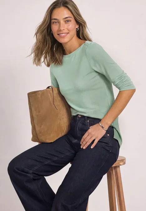 Frau sitzt auf Hocker, trägt hellgrünes 3/4-Arm-Shirt, weite Jeans und hält braune Wildledertasche.