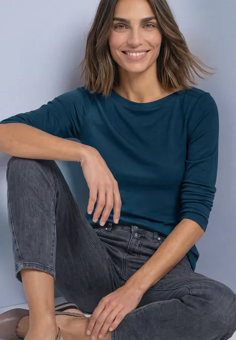 Frau in langärmeligem, dunkelblauem Oberteil und Jeans sitzt lächelnd mit einem angewinkelten Bein.
