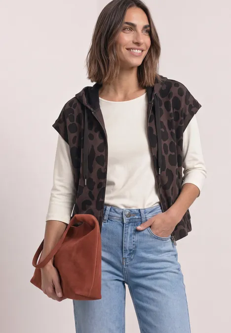 Frau in Jeans, weißem Langarmshirt, Leopardenweste mit Kapuze und brauner Wildledertasche, lächelnd.