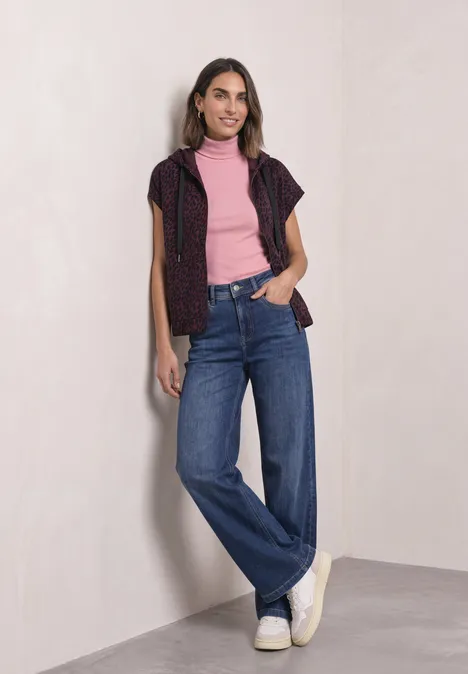 Frau lehnt lächelnd an einer Wand, trägt weite Jeans, rosa Shirt und dunklen Pullunder.