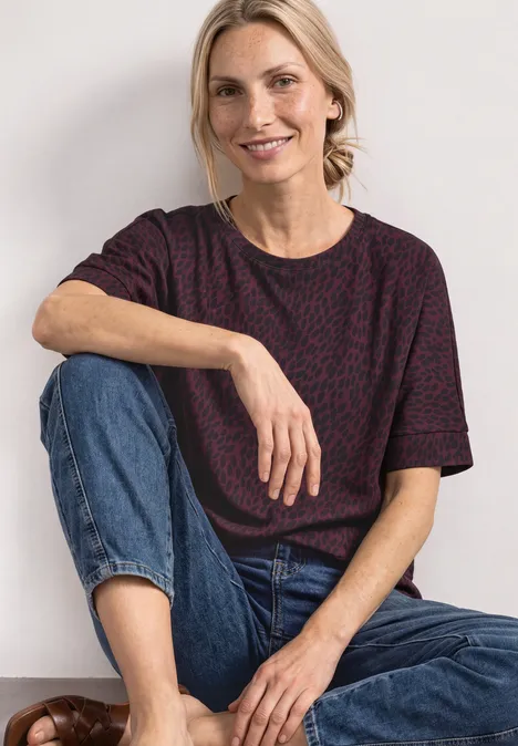 Batwing-shirt met ronde hals en print rustic rouge Een glimlachende vrouw zit ontspannen in een jeans en een gemusterd t-shirt tegen een lichte achtergrond.