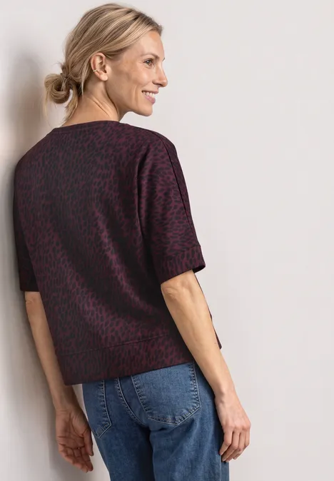 Batwing-shirt met ronde hals en print rustic rouge Een glimlachende vrouw in spijkerbroek en kortemouwenblouse met motief leunt tegen een lichte muur.