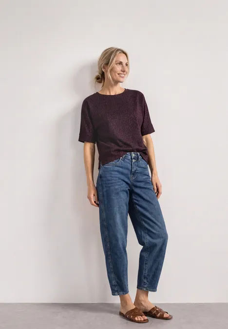 Batwing-shirt met ronde hals en print rustic rouge Een glimlachende vrouw leunend tegen een lichte muur, in een paarse top, hooggetaillede opgerolde jeans en bruine sandalen.