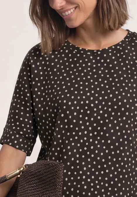 Batwing-shirt met ronde hals en print black coffee Vrouw in zwart blouse met witte stippen, houdt een bruine gevlochten tas vast.