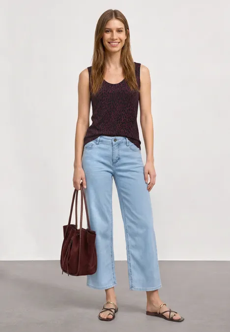 Een vrouw draagt een donker topje, een lichte jeans, sandalen en houdt een rode tas vast.