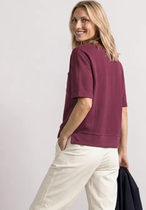 T-Shirt mit Rundhals und Struktur dark clouded blush Frau mit weinrotem Kurzarm-Sweatshirt und hellen Hosen blickt lächelnd über die Schulter.