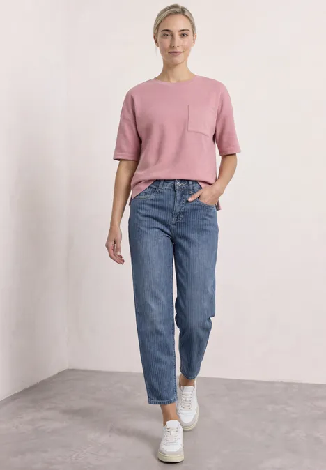 Shirt met ronde hals en structuur vintage blush Vrouw in een roze T-shirt, blauwe jeans en witte sneakers staand voor een lichte muur, ontspannen en glimlachend.