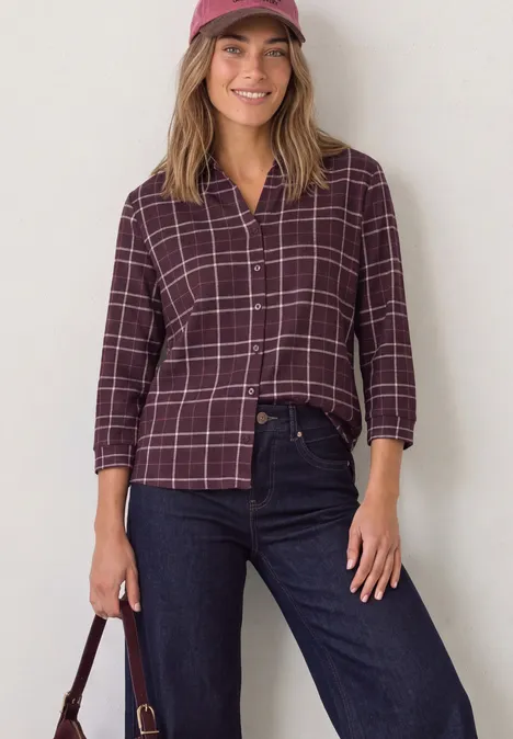 Shirt met 3/4-mouwen van jersey in blouse-stijl rustic rouge Jonge vrouw glimlacht, draagt een bordeaux geruite blouse, donkere jeans, pet en houdt een tas vast.
